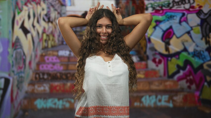 Fototapeta premium Woman smiling with hands behind head and fingers making bunny ears in a street graffiti stairwell; playful.