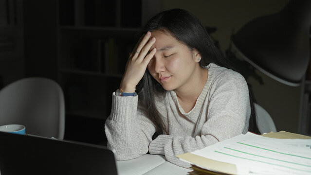 Woman student with hand on forehead over laptop and paperwork in a building study area; stress exhaustion study.