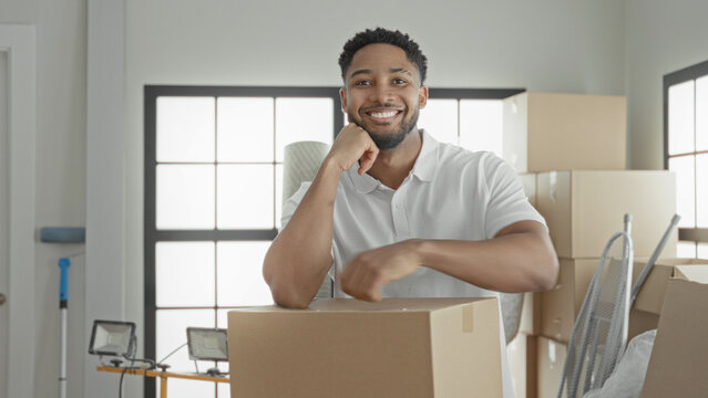 Smiling man holds a cardboard box, rests arm on it and raises fist beside stacked boxes and floodlight in building; moving day confidence.