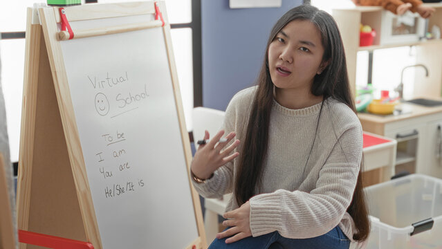 Woman teacher gestures with marker at whiteboard in studio, pointing hand visible while explaining present tense verbs to camera; engaged learning connection.