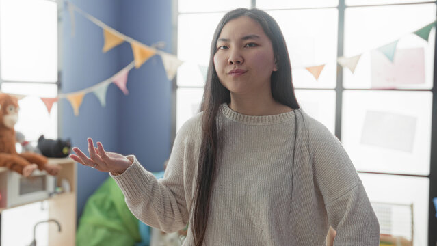 Young woman gesturing with open hand beside stuffed monkey and colorful bunting in a classroom play area, interacting with toys; mild frustration.