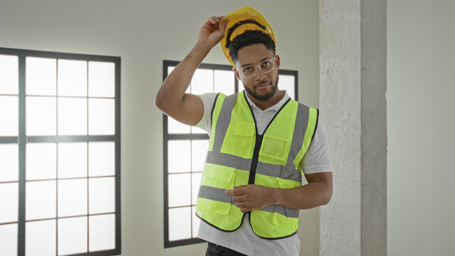 Man engineer holding a yellow hardhat and wearing a safety vest in a building with concrete column and large window; confidence.