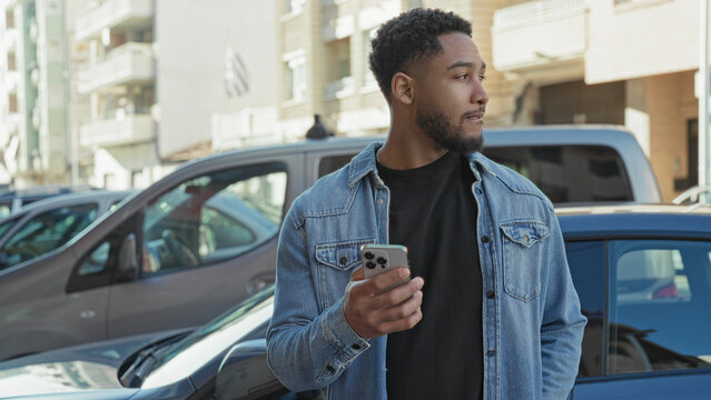 Man holding smartphone in right hand while glancing sideways near parked cars by apartment buildings on a street; thoughtful urban pause.