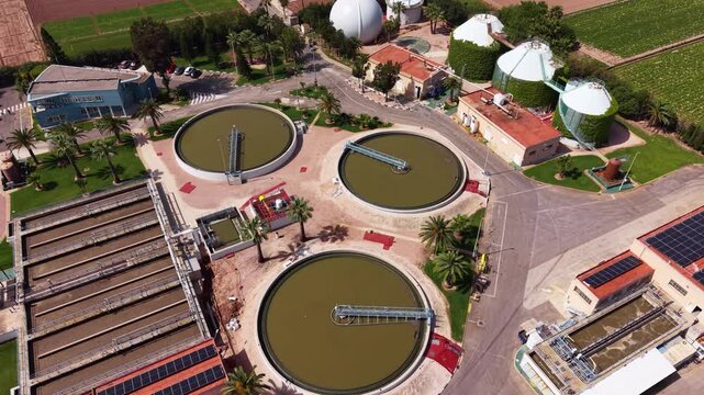 Overhead view of three circular clarifier tanks with rotating skimmer arms at a wastewater treatment facility, surrounded by palm trees, solar panels, and digester tanks