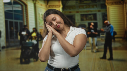 Fototapeta premium Woman resting head on hands, eyes closed, hands to cheek gesture in ornate building lobby with stroller and travelers in soft light; calm rest.