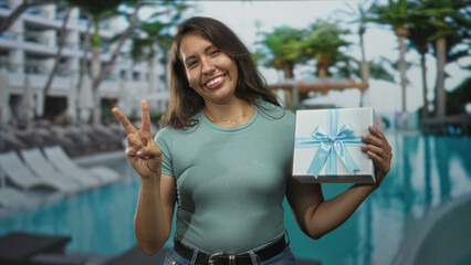 Fototapeta premium Woman in teal shirt holding wrapped gift box by pool makes peace sign and smiles with casual pose; celebration joy.