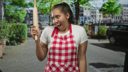 Fototapeta premium Woman cook smiling with eyes closed holds rolling pin upright on city street wearing red checkered apron and white tee; culinary joy.