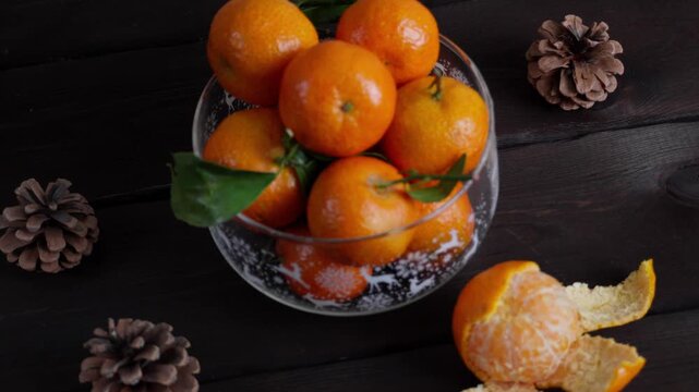 Glass Christmas bowl filled with fresh tangerines on a dark wooden table. Peeled mandarin and pine cones create a cozy winter holiday atmosphere. Festive seasonal food background