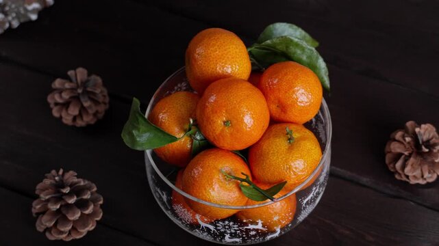 Glass Christmas bowl filled with fresh tangerines on a dark wooden table. Peeled mandarin and pine cones create a cozy winter holiday atmosphere. Festive seasonal food background