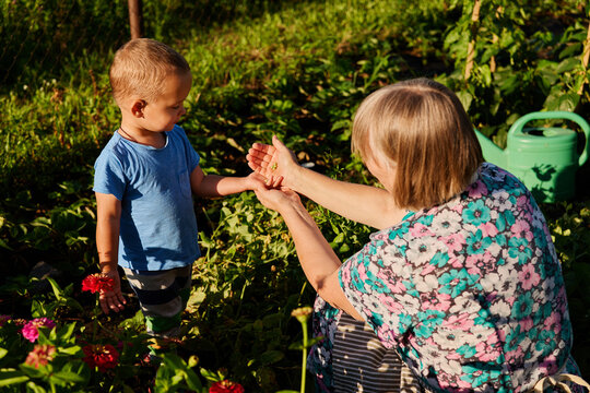 An elderly woman shows a young boy how to care for plants in a backyard garden during the late afternoon. They are surrounded by greenery and flowers