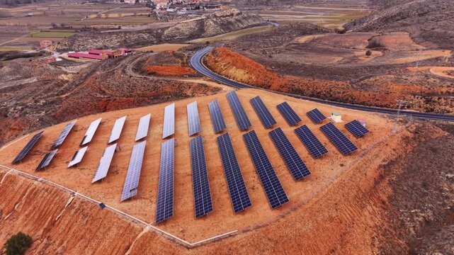 Ground-mounted solar panel installation on eroded red-earth hillside in Teruel, Aragon, Spain, with a small village, patchwork farmland, and winding road in the background