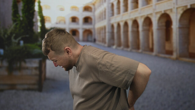 Man holding lower back and bending forward in an old town building courtyard, backache; discomfort fatigue.
