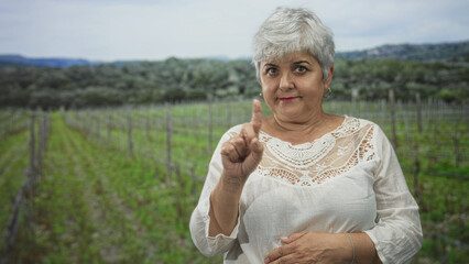Obraz premium Woman, senior with grey hair, points finger and holds hand on stomach in a vineyard forest, wearing white lace blouse and hoop earring; firm admonition.