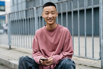 Obraz premium Young man seated by metal railing holding smartphone for casual portrait banner and social media background in urban public space