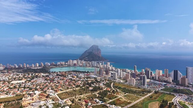 Panoramic aerial view of Calpe, Spain, showing high-rise buildings, turquoise salt lagoon, and the iconic Penon de Ifach limestone rock rising from the Mediterranean Sea