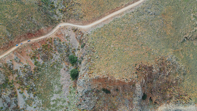 Top down aerial view of tourist cars driving on winding dirt road along steep mountain rocky slopes of Greece Mediterranean region in summer