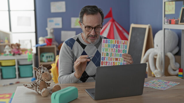 Man holding colorful alphabet chart and pointing with pen at letters beside laptop in school building; teaching focus.