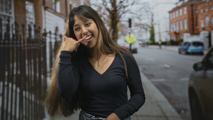 Fototapeta premium Woman with hand to ear in phone call gesture smiling by an iron fence and parked car on a street; playful invitation.