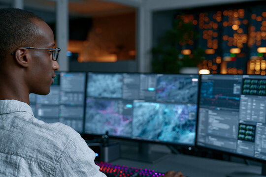 Man watches data streams across multiple monitors. His glasses reflect the glow of screens behind him. Fingers rest near a glowing keyboard, ready to act. City lights shine softly through the window