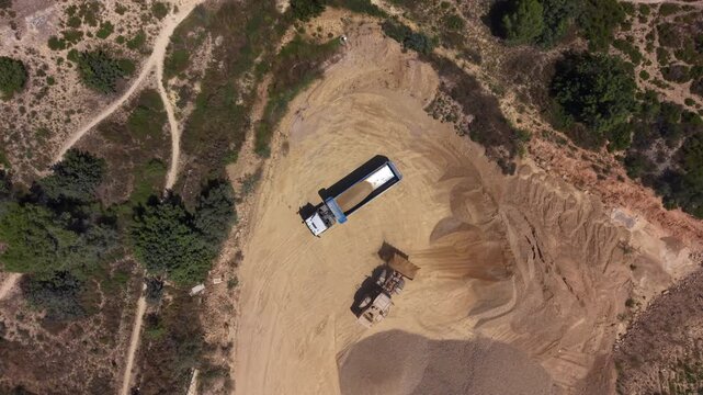 Top-down aerial view of a dump truck unloading sand and gravel onto a stockpile while a wheel loader works nearby at an open-pit aggregate quarry surrounded by trees