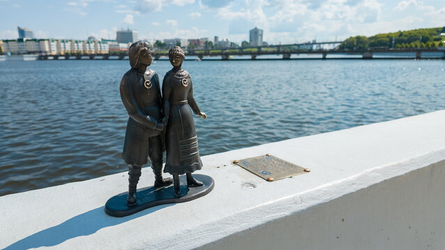 Cheboksary, Russia - June 13, 2026: A couple of bronze figurines stands by the water in Cheboksary. These mini-sculptures represent characters from Chuvash myths. The city skyline is visible.