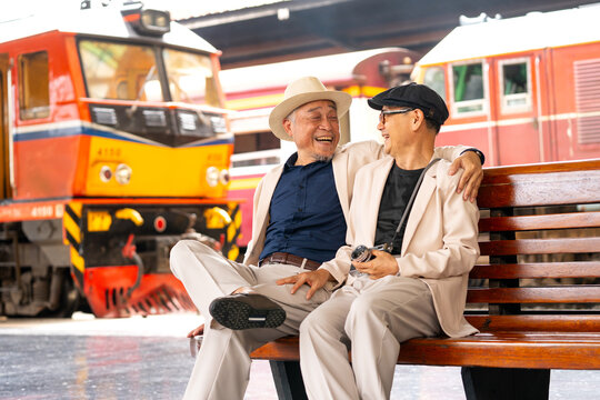 Couple Asian senior lover of lgbtq sit on long bench in train station in front of train locomotives with happiness together and one man hug to the other one.