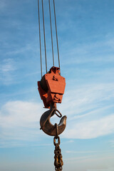 Fototapeta premium Close-up of a heavy-duty construction crane hook suspended on steel cables against a blue sky with light clouds. Industrial lifting equipment used on construction sites. Vertical photo.