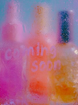 Blurred colorful serum bottle shapes behind a fogged glass surface with the words coming soon visible, creating a vibrant and intriguing visual effect