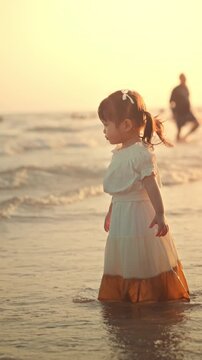 Cute little Asian girl in dress playing with sea water on tropical beach at sunset, happy curious child enjoying summer vacation and exploring nature during golden hour for family travel and parenting