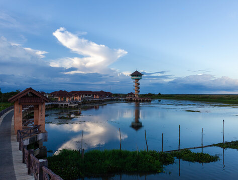 Thale Noi Viewpoint Tower overlooking a serene lake in Thailand. Walkway extends along the water's edge. The sky is clear with scattered clouds, reflecting on the calm water surface.