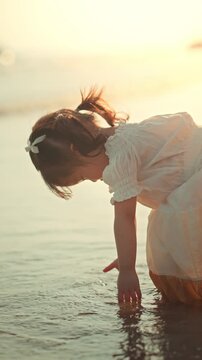 Cute little Asian girl in dress playing with sea water on tropical beach at sunset, happy curious child enjoying summer vacation and exploring nature during golden hour for family travel and parenting