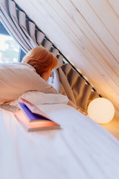 Vertical red-haired woman relaxing with books in cozy sleeping loft of modern wooden cabin