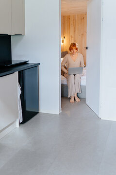 Vertical mature red-haired woman working remotely on laptop from cozy modern cabin bedroom