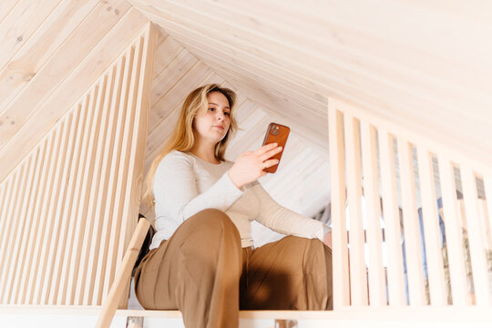 Young woman using smartphone while relaxing in bright wooden sleeping loft of modern tiny cabin