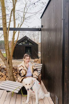 Vertical young woman enjoying hot drink with golden retriever outside modern black tiny cabin