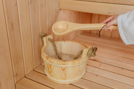 Close-up of woman pouring water from wooden sauna ladle into traditional bucket on bench