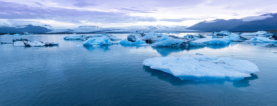 Panoramic view of J&ouml;kuls&aacute;rl&oacute;n Glacier Lagoon in southeast Iceland, where floating blue icebergs drift across calm waters beneath Vatnaj&ouml;kull Glacier and rugged mountain peaks.