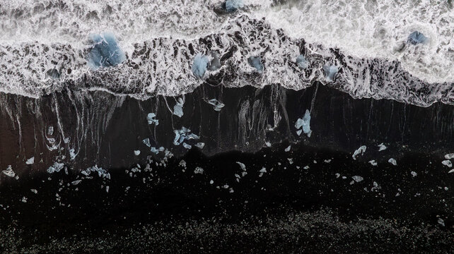 Aerial view of glacier ice fragments scattered across the black sands of Diamond Beach in Iceland, where powerful ocean waves create striking natural patterns and dramatic Arctic contrast.