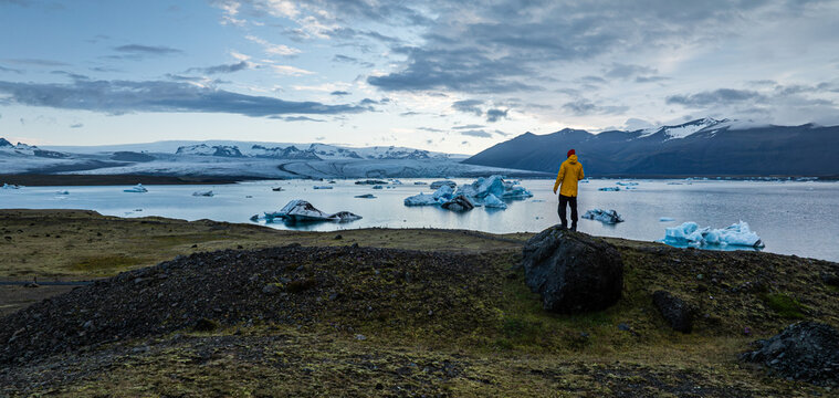Traveler standing above J&ouml;kuls&aacute;rl&oacute;n Glacier Lagoon in southeast Iceland, overlooking floating icebergs, Vatnaj&ouml;kull Glacier and rugged mountains beneath a dramatic evening sky.