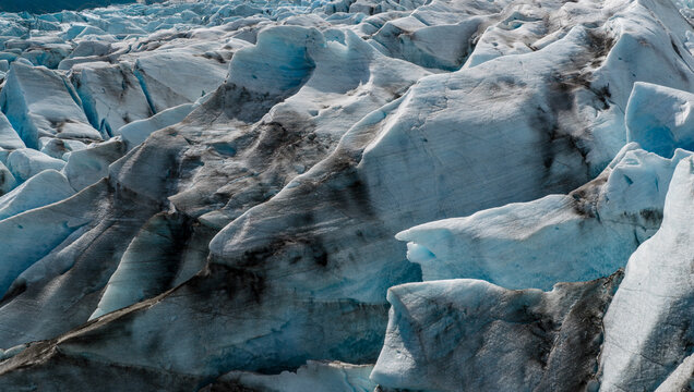 Close-up aerial view of textured glacier ice in Iceland, revealing sculpted formations, deep blue crevasses, and volcanic ash layers. A striking abstract pattern created by nature.