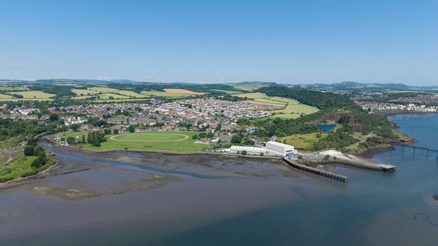 Wide angle aerial panorama of Inverkeithing coastal village in Fife, Scotland, featuring the muddy coastline at low tide, industrial piers, green fields, and residential town under a clear blue sky