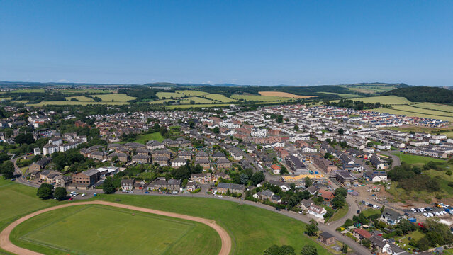 Inverkeithing, Fife, seaside village aerial drone view with residential houses, green hills, fields, and sports ground under a clear sky in Scotland