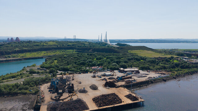 Aerial drone view of Prestonhill Quarry and an industrial metal scrap yard in Inverkeithing, Fife, with the three Forth bridges spanning the estuary in the background under a clear blue sky in Scotlan