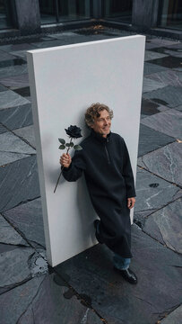 Man holding black rose against white monolithic slab in outdoor courtyard with wet stone pavement under diffused daylight, wearing long dark garment and denim trousers.
