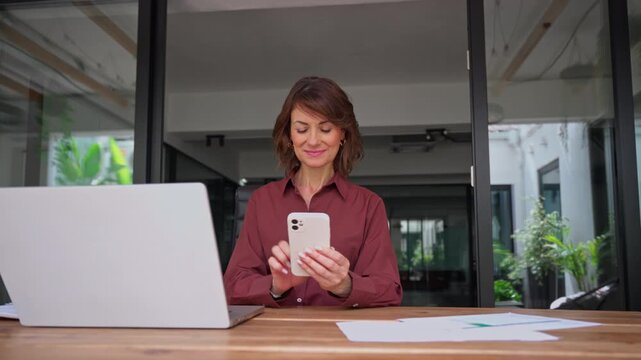 Smiling latin businesswoman holds smartphone at desk in office with copy space. Mature woman marketing director checks mobile phone app, posts business content in social media at workplace.