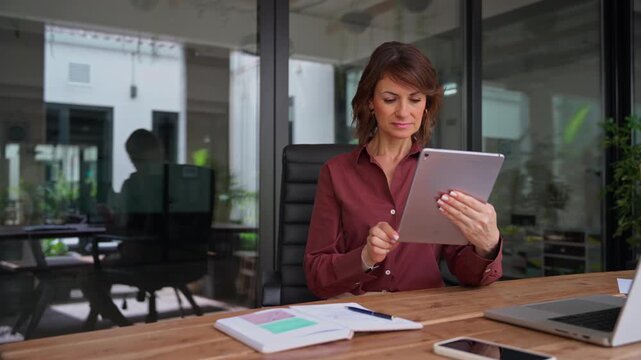 Working on digital tablet in modern office, latin mature businesswoman reads report online. Focused woman startup founder using device at desk. Female owner grows business company at workplace.