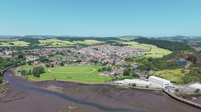 Aerial view of the coastal village of Inverkeithing in Fife, Scotland, flying towards the residential area, green sports field, and muddy bay during a bright summer day