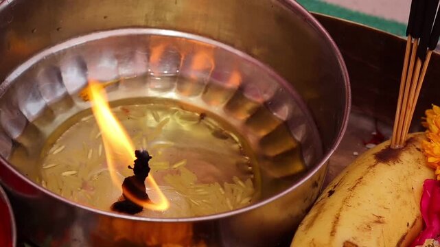 Close-up of a brightly burning traditional oil lamp with rice grains in a metal bowl on a Hindu puja thali, surrounded by incense sticks and religious offerings.
