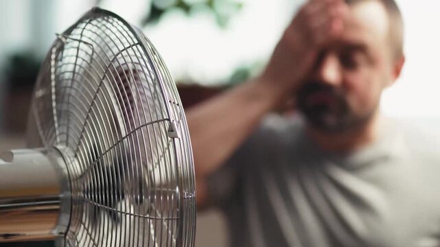 Exhausted man suffering from headache near fan in heatwave