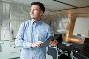 Fototapeta premium Man in a light blue shirt stands thoughtfully in a modern office while using a tablet